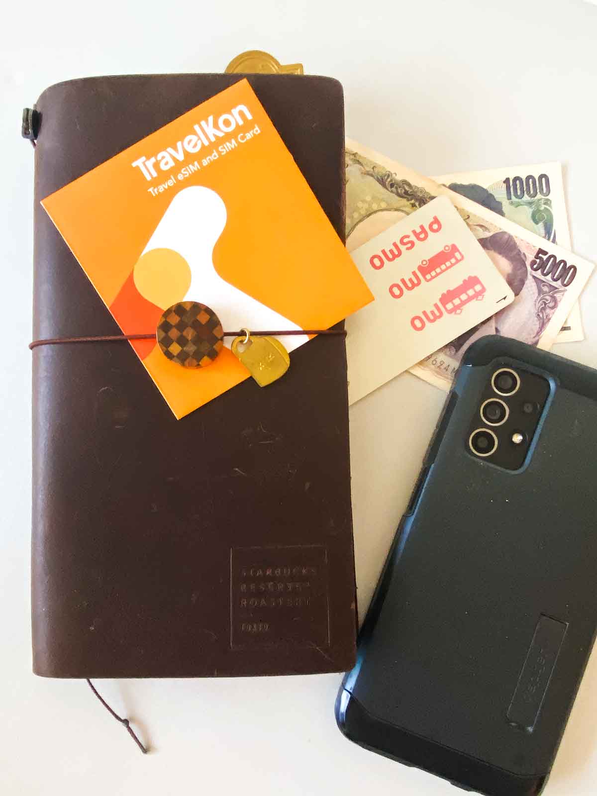 Essentials in my bag when travelling in Japan, shows Travelers Notebook, cash, IC card, TravelKon packaging and phone